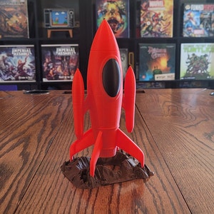 May include: A bright red rocket ship model with black oval window details and fins, standing on a brown, textured base. The rocket is displayed on a wooden table, suggesting a decorative or collectible item.