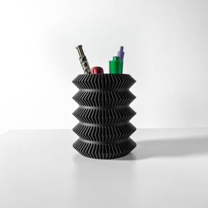Può includere: Organizzatore da scrivania geometrico nero con un motivo a zig-zag. Contiene una penna argentata, una penna verde e una penna rossa.