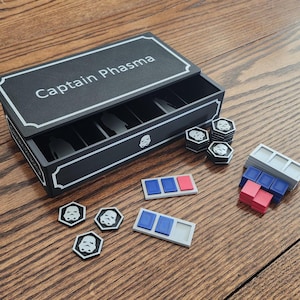 May include: A black box with the text "Captain Phasma" in white, along with various game pieces. The box has a white border and a Stormtrooper helmet icon. Several hexagonal tokens and rectangular pieces are scattered around the box.