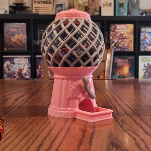May include: A pink and gray tabletop dice tower with a gumball machine design. The tower has a crank on the side and a tray at the bottom to catch the dice. The tower is filled with various colored dice.