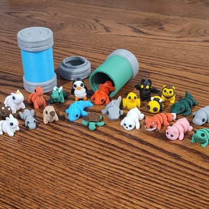 May include: A collection of small, colorful, 3D printed animal figurines, including a unicorn, a penguin, a turtle, a frog, a fox, and a bee. The figurines are arranged on a wooden surface next to two blue and green containers.