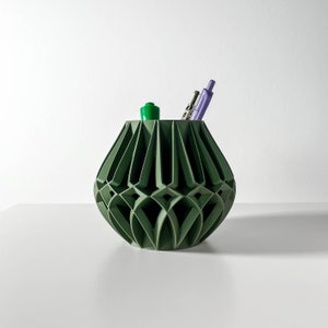 Può includere: Un vaso in ceramica verde geometrico con un design unico e intricato. Il vaso contiene due utensili da scrittura, uno verde e uno viola.