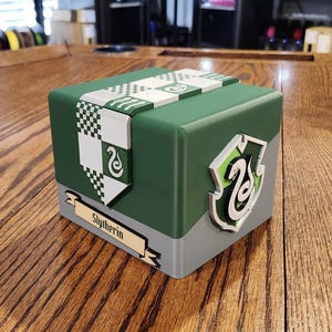 May include: A square green and gray storage box featuring a white banner with a checkered pattern and a snake emblem. A raised shield with a snake icon is attached to the side, and a beige plaque with the text Slytherin is centered below.