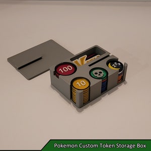 May include: A grey plastic box with compartments for storing game tokens. The box has a lid that lifts off to reveal the compartments. The compartments are filled with different coloured tokens, including red, yellow, green, and blue. The tokens have different numbers and symbols on them, including 10, 100, a skull, and a crescent moon with the letters ZZZ.