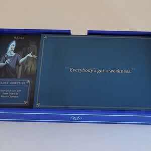 May include: A blue game board with a card featuring Hades, the Greek god of the underworld. The card has a quote: "Everybody's got a weakness."