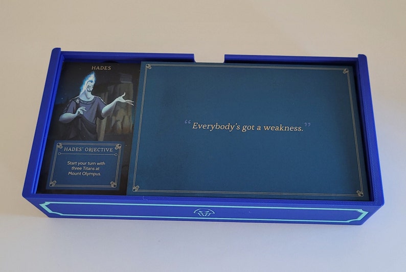 May include: A blue game board with a card featuring Hades, the Greek god of the underworld. The card has a quote: "Everybody's got a weakness."