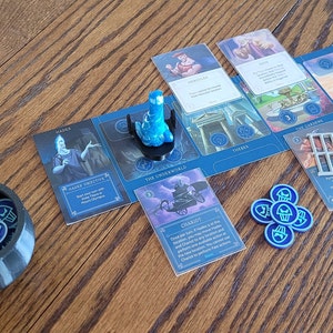 May include: A blue and white board game with a blue plastic game piece, a black cauldron filled with blue tokens, and several cards with illustrations and text. The cards include "Hades", "Power Creative", "The Underworld", "Chariot", "The Cradles", "Three Sisters", and "Mount Olympus".