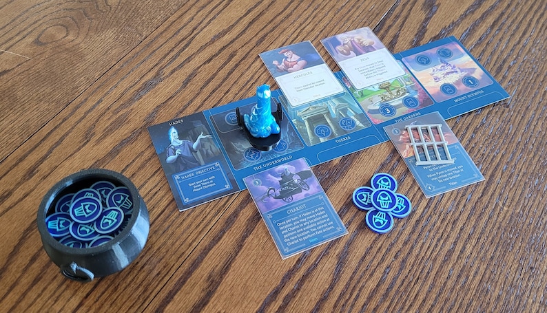 May include: A blue and white board game with a blue plastic game piece, a black cauldron filled with blue tokens, and several cards with illustrations and text. The cards include "Hades", "Power Creative", "The Underworld", "Chariot", "The Cradles", "Three Sisters", and "Mount Olympus".