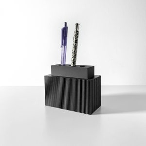 Può includere: Un portapenne nero con una texture a coste e due penne al suo interno. Una penna è viola e l'altra è nera e bianca.