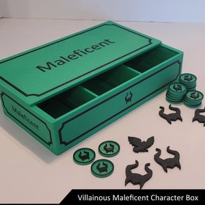 Puede incluir: Una caja verde con la palabra "Maléfica" en la tapa. La caja tiene un compartimento para guardar las piezas del juego. La caja está rodeada de piezas de juego negras con un diseño de dragón.