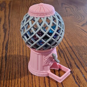 May include: A pink gumball machine with a gray wire cage filled with colorful dice. The machine has a crank on the side and a small tray underneath.