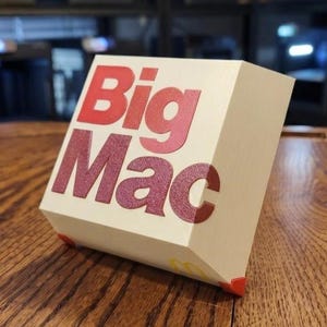 May include: A cream-colored, cube-shaped decorative object featuring the red text Big Mac on the front face. The object has a smooth, matte finish and is supported by small red corner feet. A small yellow McDonald's logo is visible on the bottom edge.