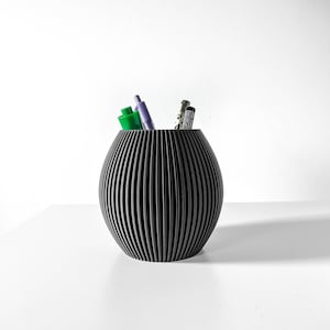 Può includere: Un portapenne nero cilindrico con un design a coste. È pieno di penne e matite, tra cui una penna verde e una penna viola.