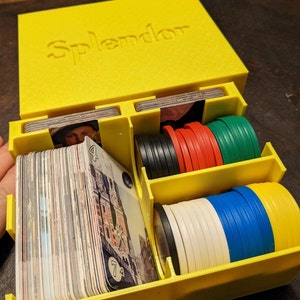 May include: A yellow plastic box with the word "Splendor" printed on the lid. The box is open and contains a variety of game pieces, including cards, chips, and tokens.