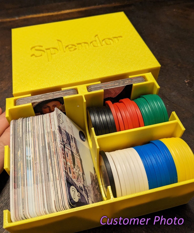 May include: A yellow plastic box with the word "Splendor" printed on the lid. The box is open and contains a variety of game pieces, including cards, chips, and tokens.