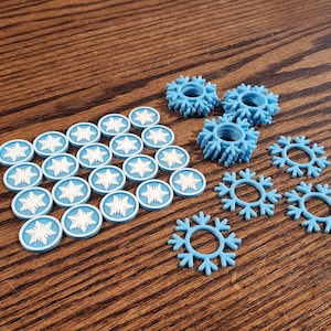 May include: A collection of light blue plastic items featuring snowflake designs. The set includes twenty circular tokens with embossed white snowflakes and seven open-ring snowflake shapes arranged in a loose cluster on the surface.