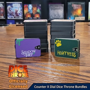 May include: Two stacks of black dial dice throne bundles with a purple and green counter on top. The purple counter has the text "Shadow Thief" and the number "50". The green counter has the text "Huntress" and the number "50". The text "Dice Throne" is visible in the bottom left corner of the image. The text "Officially Licensed" is visible below the text "Dice Throne". The text "Counter X Dial Dice Throne Bundles" is visible in the bottom right corner of the image.