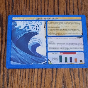 May include: A blue-bordered game card titled "Ocean's Hungry Grasp" with text and a graphic of a large ocean wave. The card includes sections for setup, play style, and a summary of powers. The complexity is marked as high.