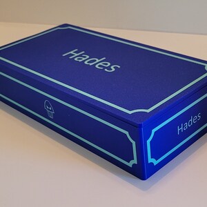 May include: A blue box with a light blue trim and the word "Hades" printed on the top and side. A small skull icon is printed on the front of the box.