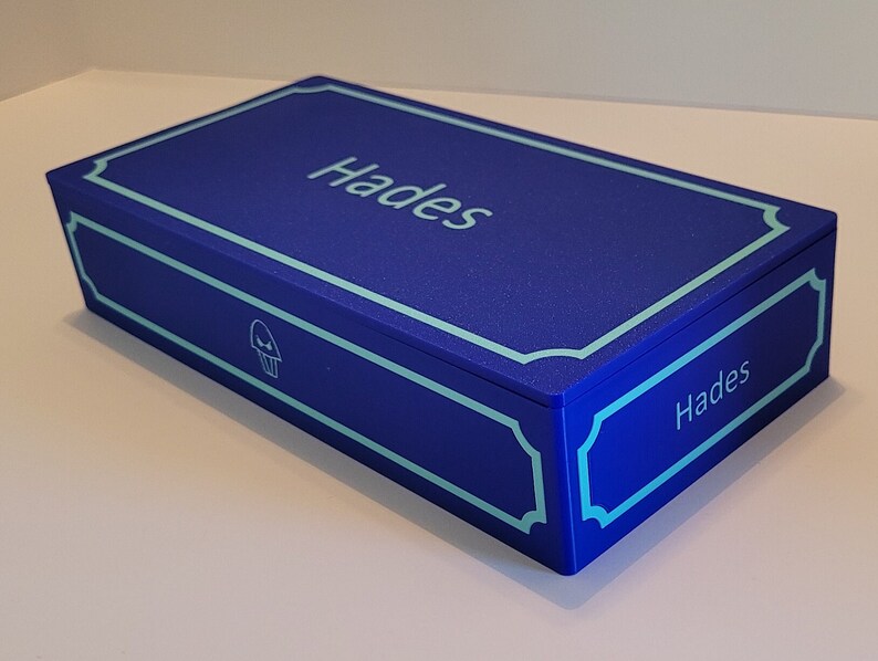 May include: A blue box with a light blue trim and the word "Hades" printed on the top and side. A small skull icon is printed on the front of the box.