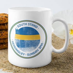 Puede incluir: Taza de cerámica blanca con un diseño circular azul y amarillo que dice "SOUTH STAND. HEADINGLEY. SUNDAY 3PM." La taza está junto a una pila de galletas con chispas de chocolate.