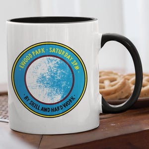 May include: A white ceramic mug with a black handle and interior. The mug features a blue and yellow circular design with the text "EWOOD PARK SATURDAY 3PM" and "BY SKILL AND HARD WORK."