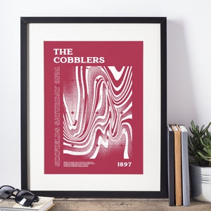 Peut inclure: Affiche encadrée avec un fond bordeaux et du texte blanc. L'affiche indique "THE COBBLERS" en haut, "SIXFIELDS SATURDAY 3PM" sur le côté gauche et "1897" en bas à droite. Le centre présente un motif ondulé blanc.