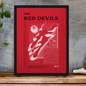 May include: Framed red and white graphic poster with the text "THE RED DEVILS". The artwork features abstract shapes and additional text. The poster is displayed on a wooden surface, with a plant in the background.