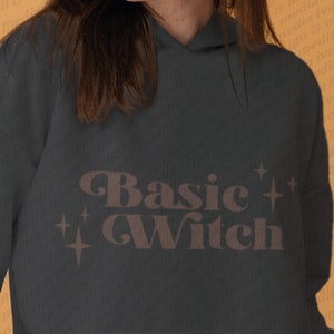 Könnte beinhalten: Ein dunkelgrauer Hoodie mit einer braunen Grafik, auf der "Basic Witch" mit zwei Sternen auf jeder Seite des Textes steht.