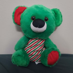 Può includere: Un orsacchiotto verde con dettagli rossi sulle orecchie e sui piedi. L'orso ha un muso bianco, un naso nero e occhi marroni. Indossa una sciarpa a righe rosse e bianche. Un giocattolo morbido e coccoloso.