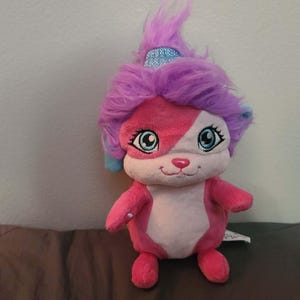 May include: A plush toy featuring a pink and white face with blue eyes and a small pink nose. The toy has a pink body, arms, and legs, and a purple head of hair. A small blue sparkly band is on the head.