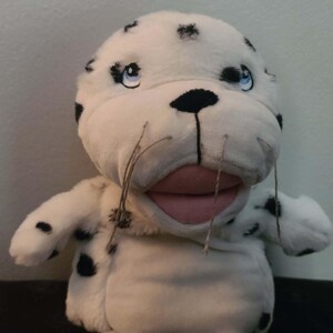 May include: A plush seal puppet with a white body and black spots. The seal has a pink mouth, blue eyes, and long whiskers. The puppet's arms are outstretched, and it appears to be smiling.