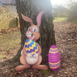 May include: A brown bunny rabbit holding a blue and yellow Easter egg. The bunny is standing next to a tree trunk. There is a purple and yellow Easter egg on the ground.