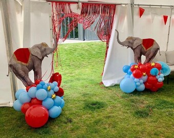 Puede incluir: Dos recortes de elefantes con mantas de silla rojas y doradas se alzan sobre arreglos de globos. Los globos son rojos y azules, con detalles de estrellas doradas. La escena está ambientada en un césped, con una carpa y flecos rojos en el fondo.