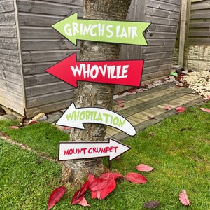 May include: Four red and white arrow signs with green and white text attached to a tree trunk. The signs point in different directions and read "Grinch's Lair", "Whoville", "Whoblation", and "Mount Crumpet".