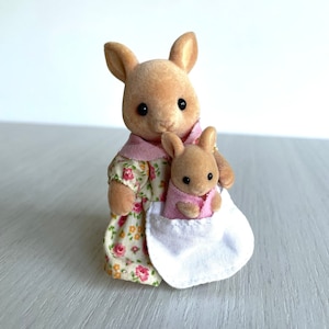 May include: A brown plush kangaroo mom figure holding a smaller brown plush baby kangaroo in a pink and white dress. The mom kangaroo is wearing a yellow and pink floral dress with a white apron.