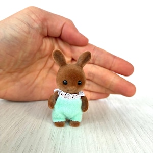 May include: A small brown rabbit figurine wearing a light green romper with a white lace collar. The toy has black eyes and is designed to look like a baby. The figurine is a collectible toy.