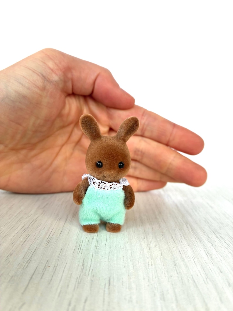 May include: A small brown rabbit figurine wearing a light green romper with a white lace collar. The toy has black eyes and is designed to look like a baby. The figurine is a collectible toy.