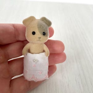 Könnte beinhalten: Kleine, hellbraune und graue Hunde-Figur mit schwarzen Augen und einem rosa-weißen Rock. Der Rock hat einen Perlenknopf und rosa Blumenstickerei. Die Ohren des Hundes sind hellbraun und grau.