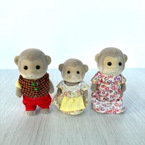 May include: Three toy monkey figures. The monkey on the left wears a yellow shirt, a brown plaid vest, and red pants. The middle monkey wears a yellow dress with a white lace collar and a yellow apron. The monkey on the right wears a floral dress.