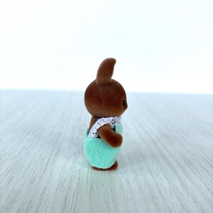 May include: A small brown bunny figurine wearing a mint green outfit with white lace trim. The bunny has a small, upright ear and is standing on a light-colored surface. The figurine is a toy or collectible.