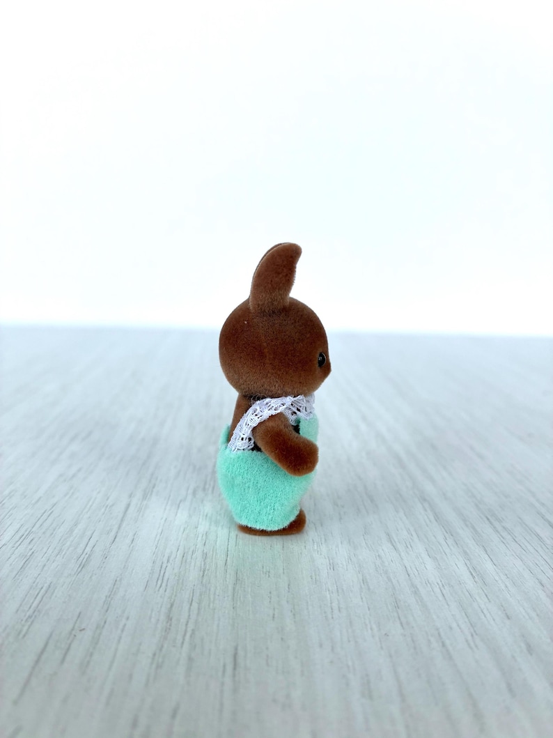 May include: A small brown bunny figurine wearing a mint green outfit with white lace trim. The bunny has a small, upright ear and is standing on a light-colored surface. The figurine is a toy or collectible.