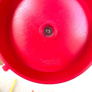 Op de afbeelding: Rode plastic basis van een speelgoedspel met een schroef in het midden. De basis heeft de tekst "EPOCH CO., LTD. MADE IN CHINA" erop gedrukt.
