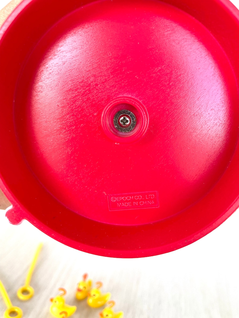 Op de afbeelding: Rode plastic basis van een speelgoedspel met een schroef in het midden. De basis heeft de tekst "EPOCH CO., LTD. MADE IN CHINA" erop gedrukt.