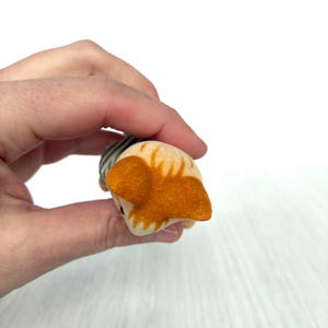 May include: A small, handmade felt cat toy in shades of tan, orange, and gray. The cat's head is tan with orange ears and markings. The body is striped gray and white. The toy is held in a hand against a white background.