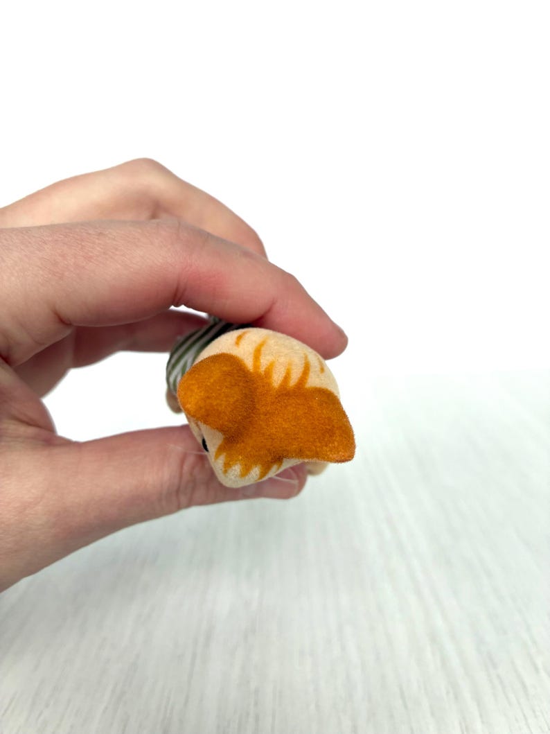 May include: A small, handmade felt cat toy in shades of tan, orange, and gray. The cat's head is tan with orange ears and markings. The body is striped gray and white. The toy is held in a hand against a white background.