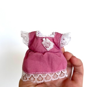 May include: A pink doll dress with white lace trim and a white button detail. The dress has a gathered skirt and short sleeves.