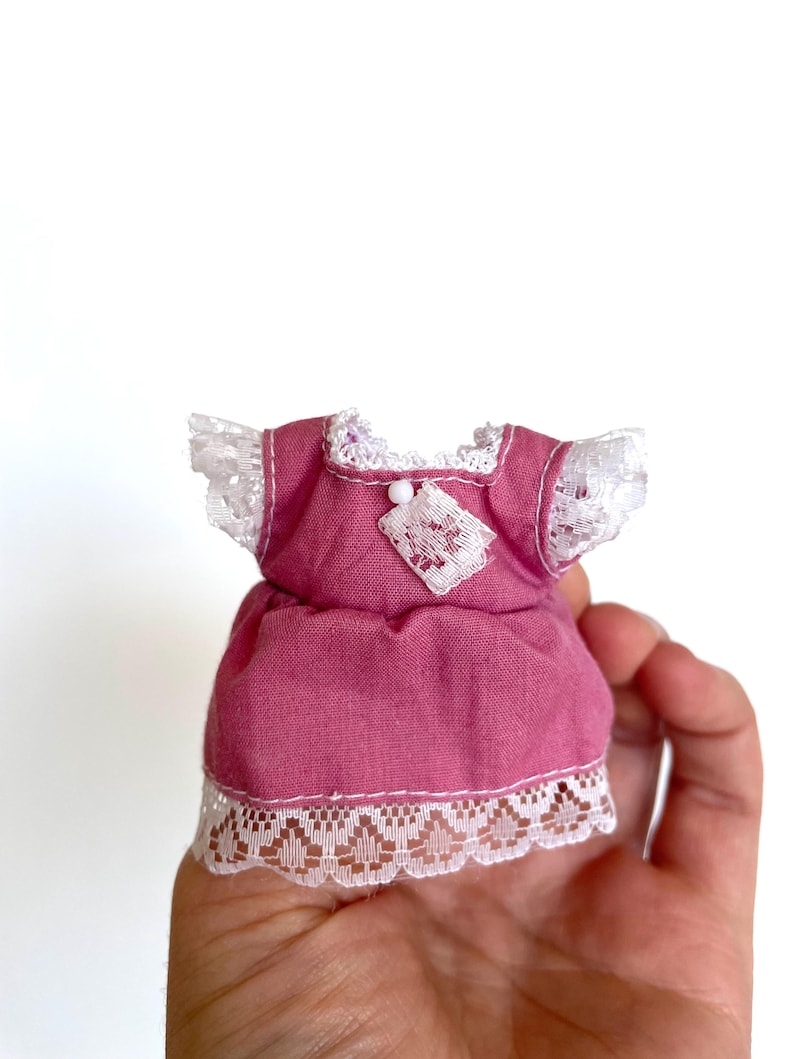 May include: A pink doll dress with white lace trim and a white button detail. The dress has a gathered skirt and short sleeves.