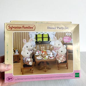Puede incluir: Una caja del juego Sylvanian Families Dinner Party. La caja presenta una escena de comedor en miniatura con figuras de conejos alrededor de una mesa con comida y bebida. La caja tiene el texto "Dinner Party Set" y "Over 40 pieces".