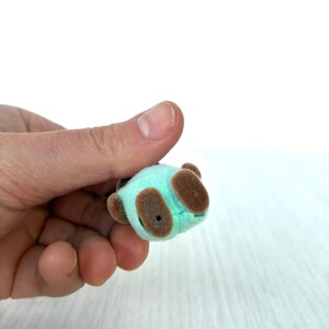 May include: A miniature plush panda bear toy in a light teal color with brown accents. The small toy has brown ears and feet. The panda is held in a person's hand against a white background.